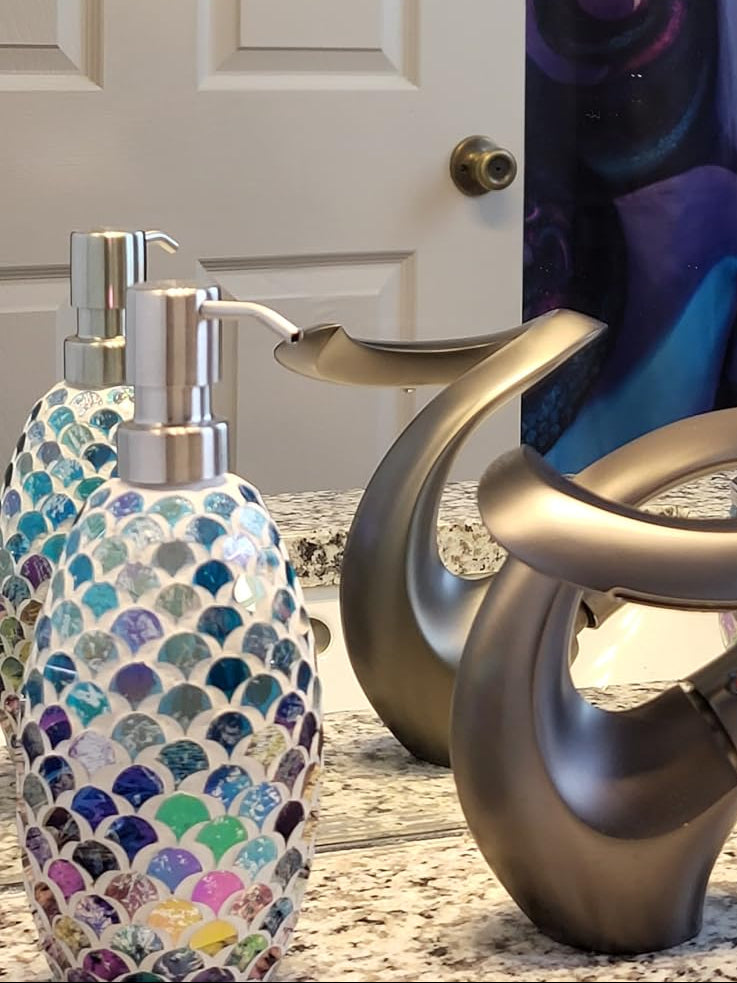 A bathroom countertop scene featuring a modern curved metallic faucet, two colorful mosaic soap and lotion dispensers, and a decorative jar, with a large Disney Ursula character print hanging on the wall behind them.